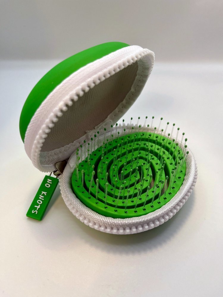 The green No Knots palm detangler sitting open inside its white lined zip case, showing the spiral bristle pad and a zip tag reading No Knots.